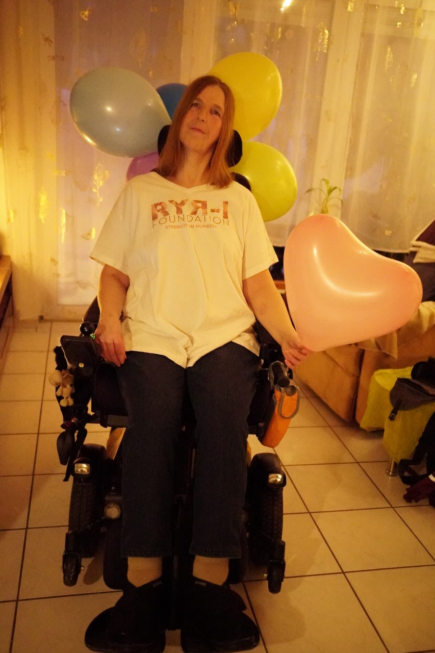 Bettina zuhause in Bramsche in ihrem Elektro-Rollstuhl. Sie trägt ein T-Shirt der RYR1 Foundation.